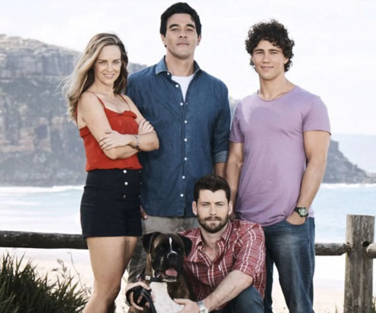 Group of four people and a dog at beachside, one sitting, others standing; scene from "Home and Away".