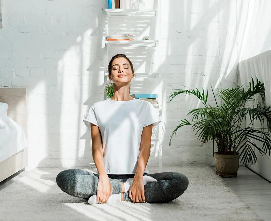 Woman meditating cross-legged on a carpeted floor in a sunlit room with plants and shelves.