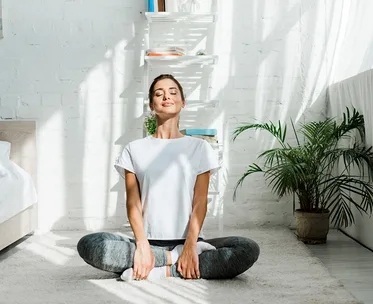 Woman meditating cross-legged on a carpeted floor in a sunlit room with plants and shelves.