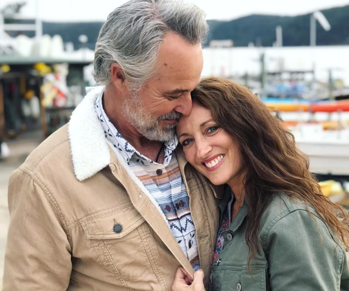 Elderly man in tan jacket embraces smiling woman in green coat by a harbor background.