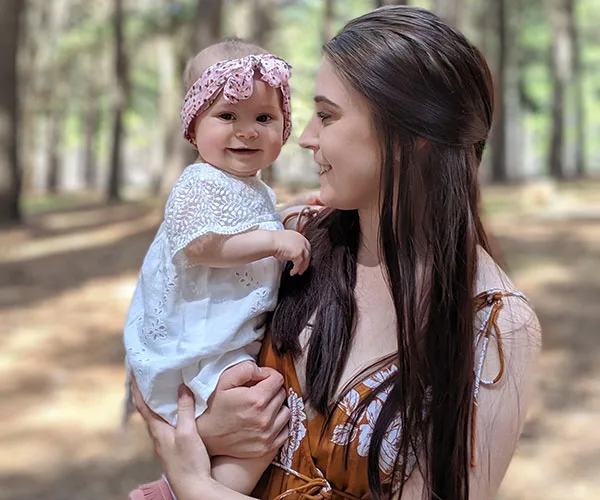 Young woman smiling at a baby wearing a pink headband, outdoors in a forest setting.