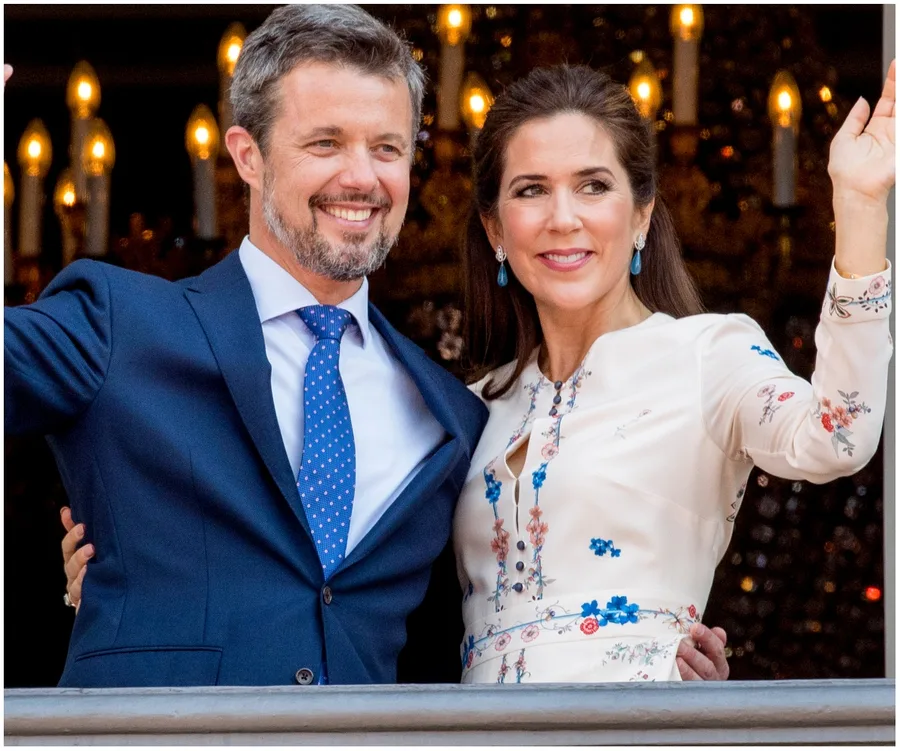Couple smiling and waving, standing together dressed in formal attire with candlelights in the background.