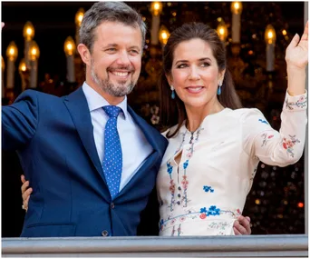 Couple smiling and waving, standing together dressed in formal attire with candlelights in the background.