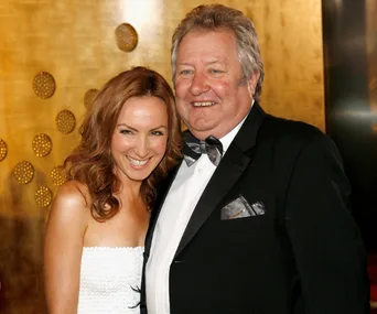 Two people smiling at a formal event, the man in a tuxedo and the woman in a white dress, against a decorative backdrop.