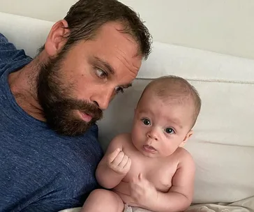 Man with beard lying on a couch, looking at a baby beside him, both appearing relaxed and comfortable.