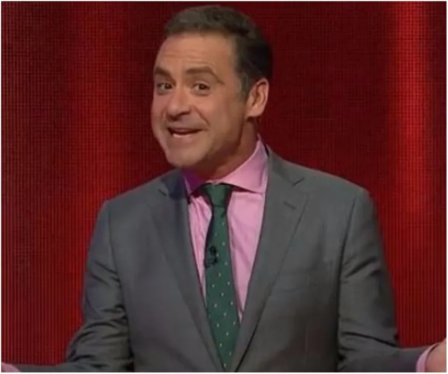 Man in a gray suit, pink shirt, and green tie, gesturing with a smile against a red background.
