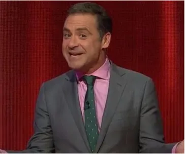 Man in a gray suit, pink shirt, and green tie, gesturing with a smile against a red background.