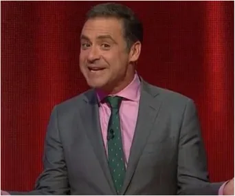 Man in a gray suit, pink shirt, and green tie, gesturing with a smile against a red background.