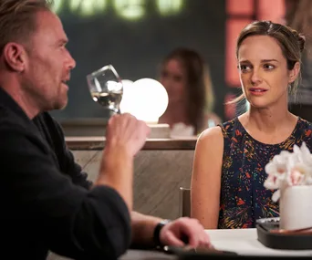 Tori and Christian from "Home and Away" on a dinner date, with Christian holding a wine glass and Tori looking intrigued.