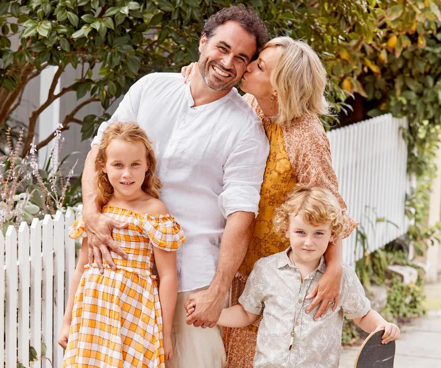 Family portrait with a smiling couple and two children, outdoors by a white fence. The boy holds a skateboard.