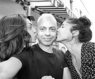 A man taking a selfie with two women kissing his cheeks, smiling in an outdoor setting.