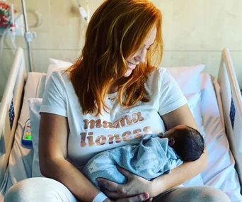 A woman with red hair, wearing a "mama lioness" shirt, smiles while holding a newborn baby in a hospital bed.