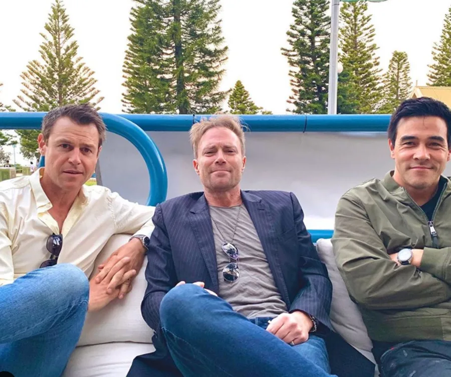 Three men sitting on an outdoor sofa, smiling, with trees in the background.