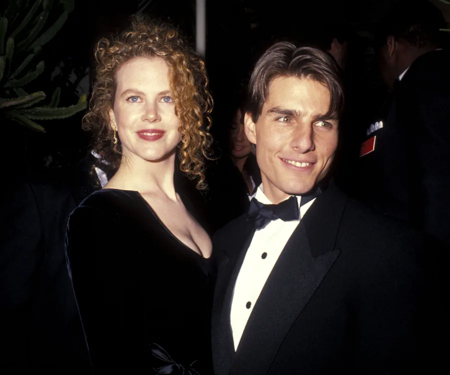 A couple in formal attire at an event, one with curly hair in a black dress and the other with slicked hair in a tuxedo.