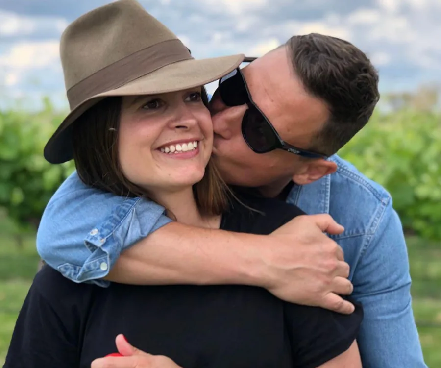 Man kisses woman on cheek, outdoors. Woman wears a hat and smiles; man wears sunglasses and blue shirt.