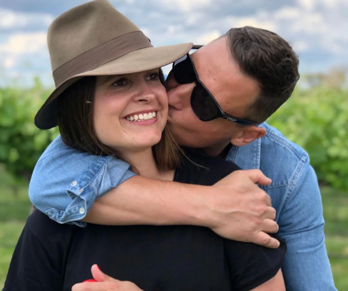 Man kisses woman on cheek, outdoors. Woman wears a hat and smiles; man wears sunglasses and blue shirt.