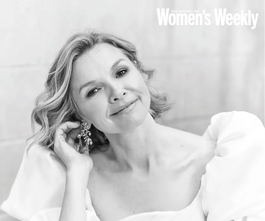 A woman with wavy hair smiles gently, wearing an elegant dress and holding a decorative earring.