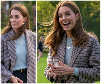 A woman in a checked blazer smiles and gestures outdoors with greenery in the background.