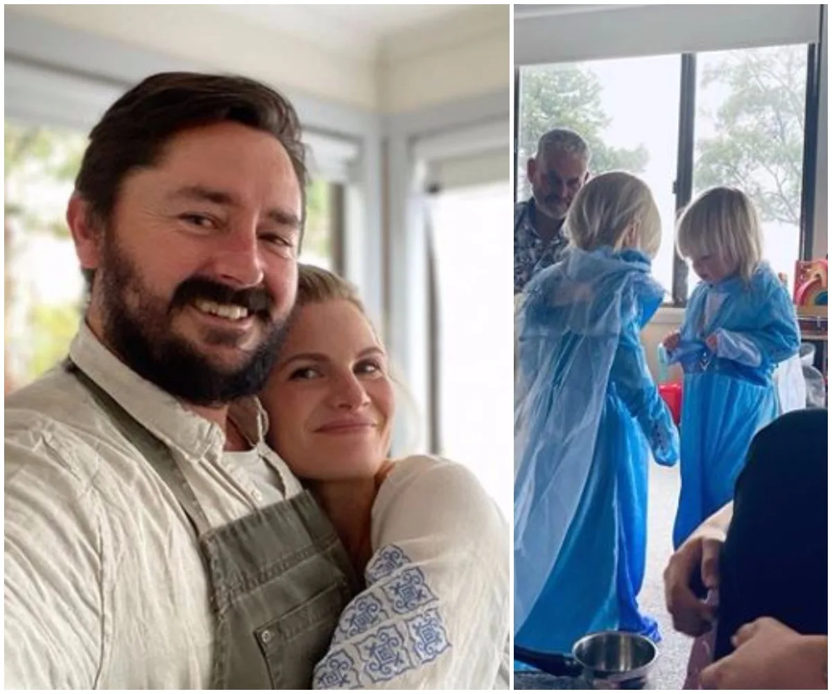 A couple smiles at the camera; two children in blue dresses play nearby indoors.