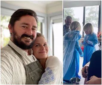 A couple smiles at the camera; two children in blue dresses play nearby indoors.