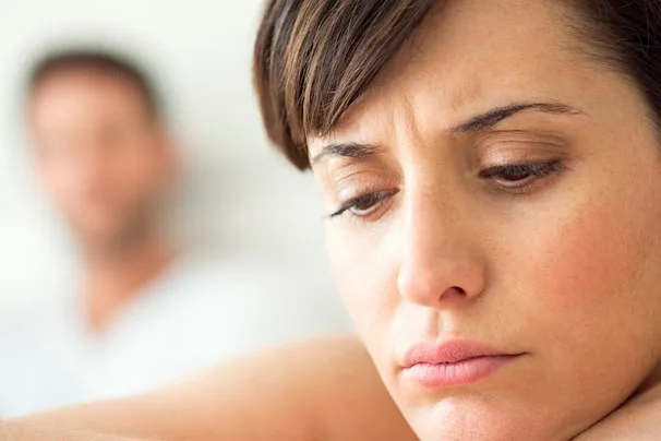 Sad woman in focus with concerned expression, blurred man in background.