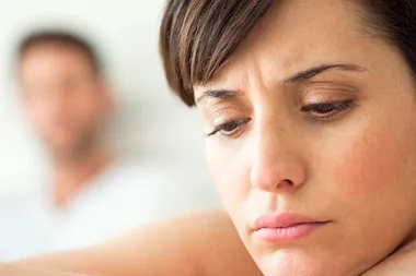 Sad woman in focus with concerned expression, blurred man in background.