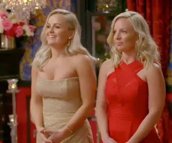 Two women in elegant dresses, one in gold and the other in red, with a background of flowers and chandeliers.