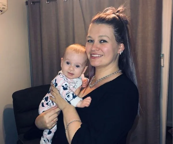 A woman with a smile holding a baby in pajamas. Brown curtains in the background.