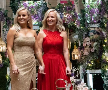 Two women smiling and holding hands, one in a gold dress and the other in red, stand amid floral decor and lanterns.