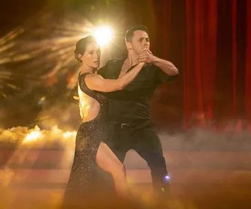 Dancers perform passionately on stage with dramatic lighting and a smoky background, wearing elegant black outfits.