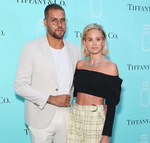 Couple posing together in front of a Tiffany & Co. backdrop; man in beige suit, woman in black top and checked pants.