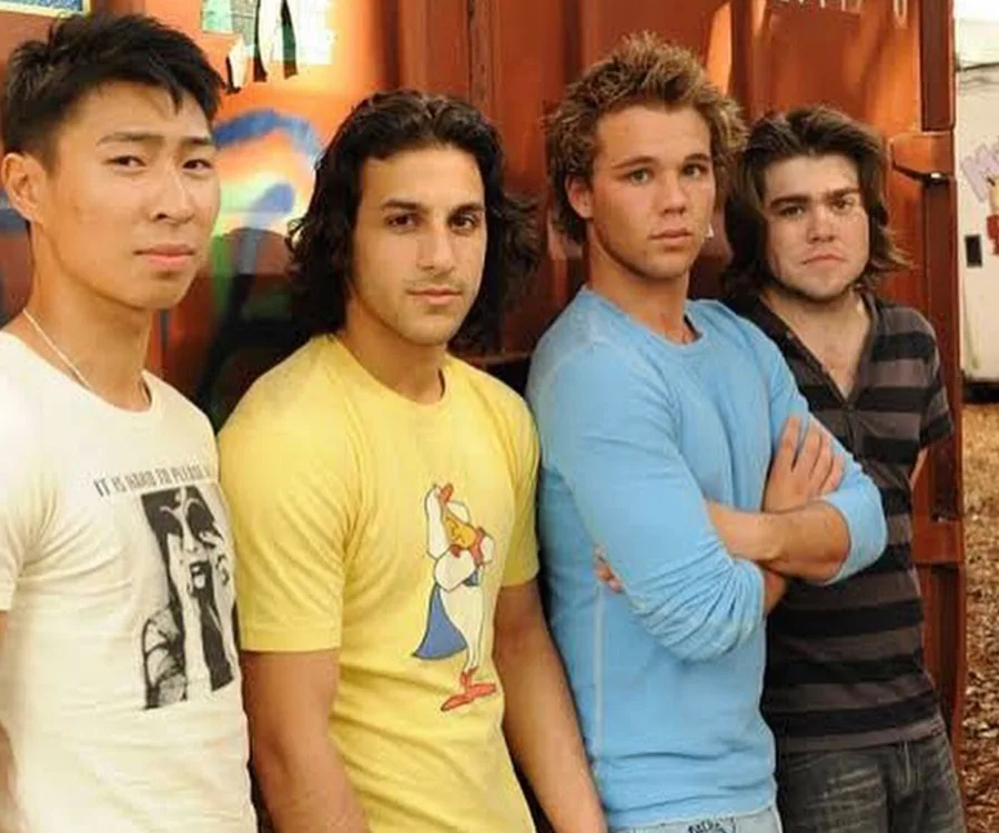 Four young men standing casually in front of a rusty, graffiti-covered container, each wearing casual shirts.