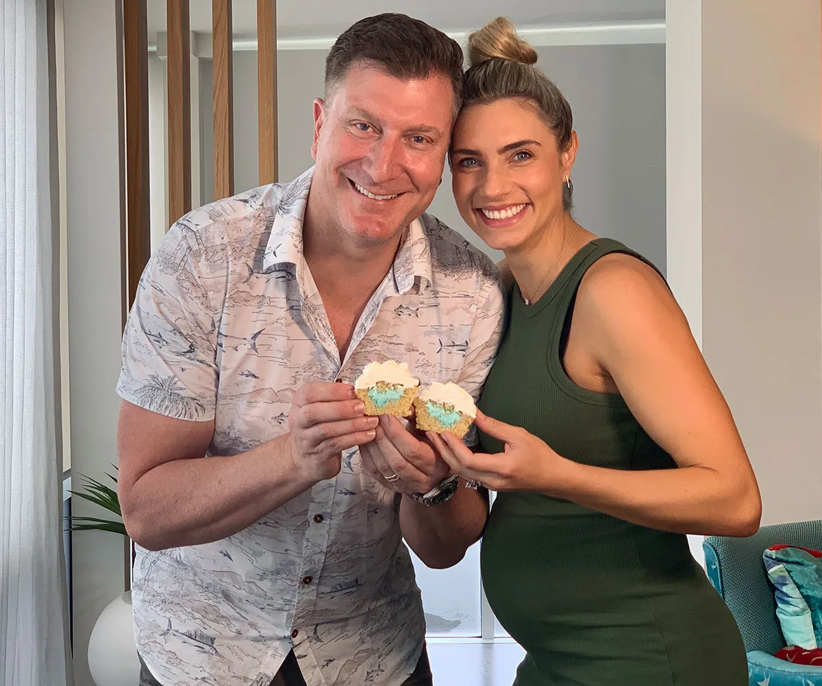 Smiling couple holding cupcakes with blue filling, suggesting a gender reveal for a baby boy, indoors.