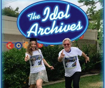 Two people in matching t-shirts pose playfully under a "The Idol Archives" sign in a garden setting.