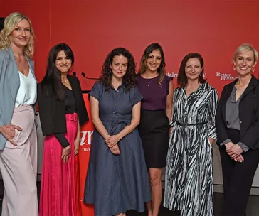 A group of six women posing together at an event against a red background with logos.