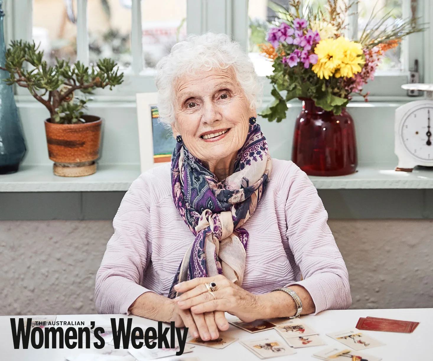Elderly woman smiling in a cozy setting with plants and cards on the table, featured in The Australian Women's Weekly.