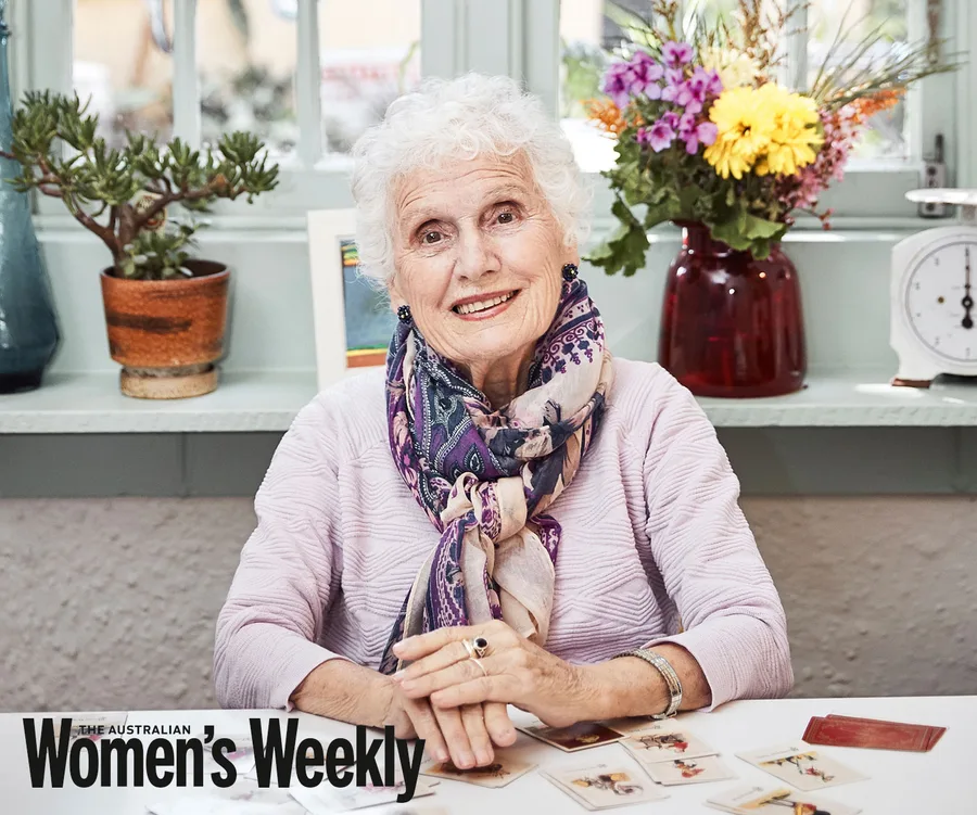 Elderly woman smiling in a cozy setting with plants and cards on the table, featured in The Australian Women's Weekly.