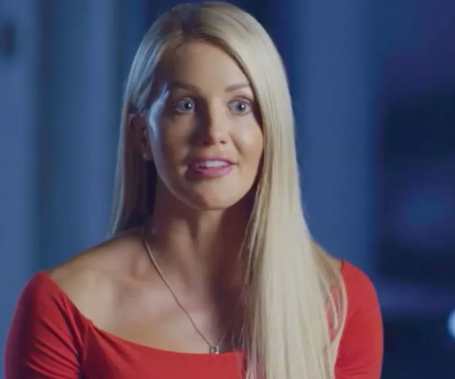 Woman with long blonde hair in a red top, speaking in a dimly lit room.