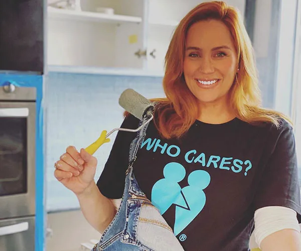 Woman smiling in a kitchen, holding a paint roller, wearing a "Who Cares?" T-shirt and denim overalls.
