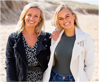 Two women smiling on a beach, one in a black floral dress and jacket, the other in a green top and white coat.