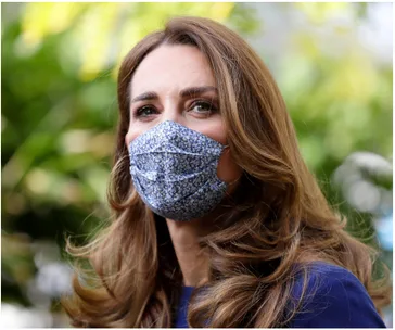 Woman with long brown hair wearing a blue floral mask, standing outdoors with a blurred green background.