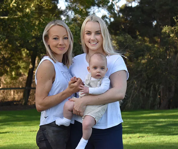 Two women smiling outdoors, one holding a baby. Trees and grass are in the background.