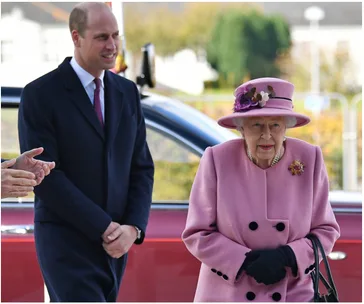 Elderly woman in pink coat and hat, standing with a younger man in a suit, near a red car.