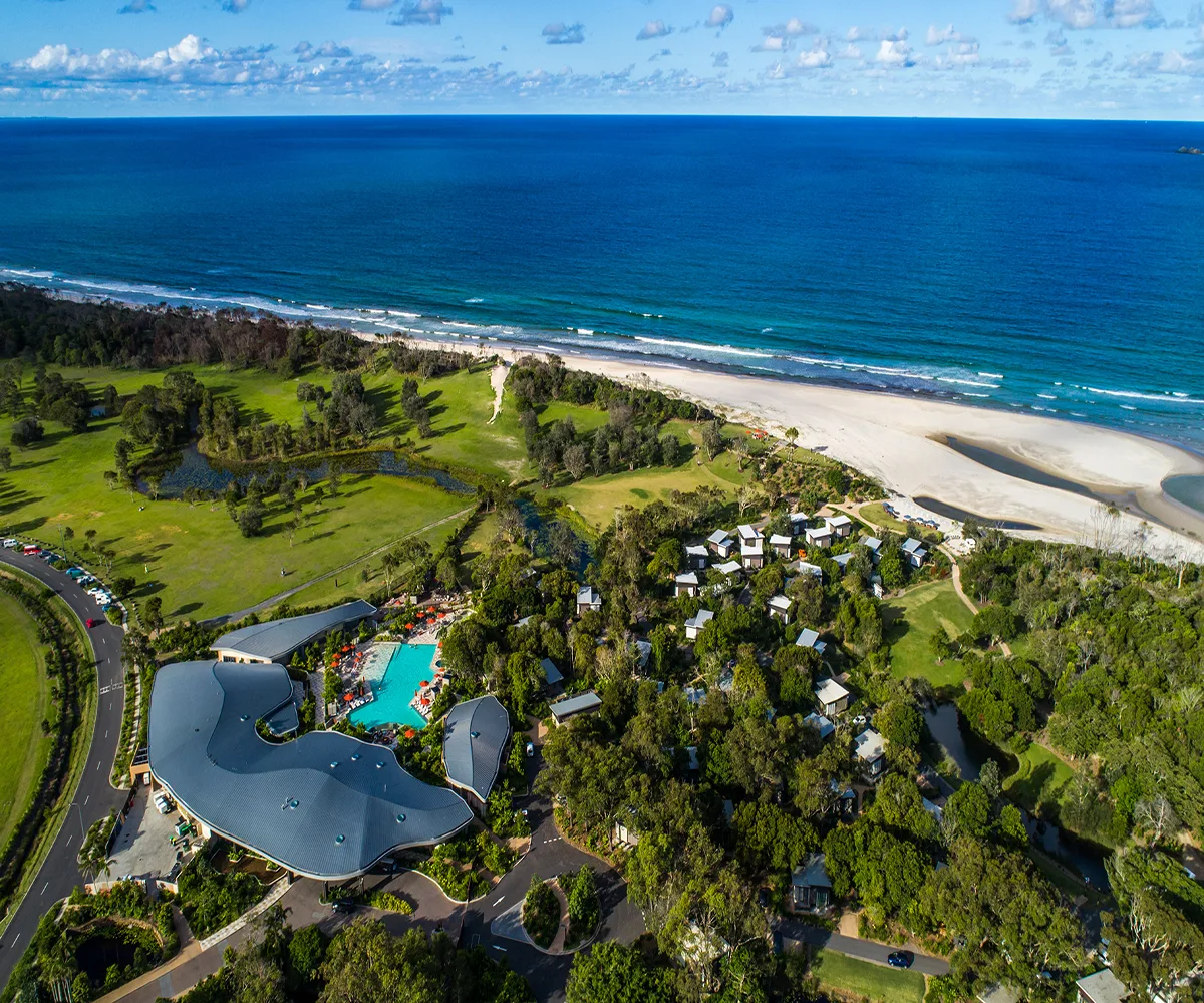 Aerial view of a resort near Byron Bay, Australia, with a pool, green landscape, and ocean in the background.