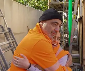 Two people in orange work attire warmly embracing at a construction site.