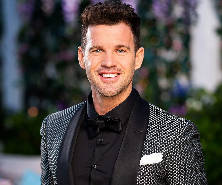 Man in tuxedo smiling outdoors, with elegant garden backdrop.
