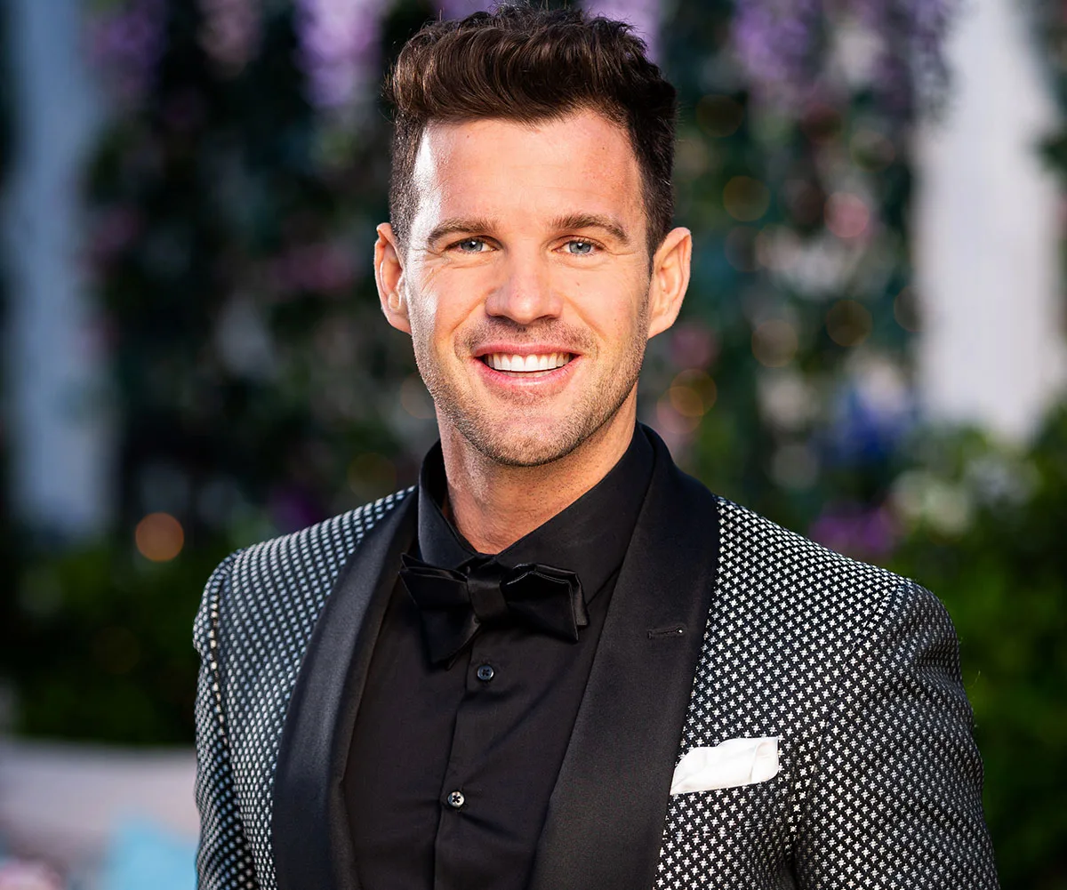 Man in tuxedo smiling outdoors, with elegant garden backdrop.