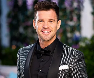 Man in tuxedo smiling outdoors, with elegant garden backdrop.