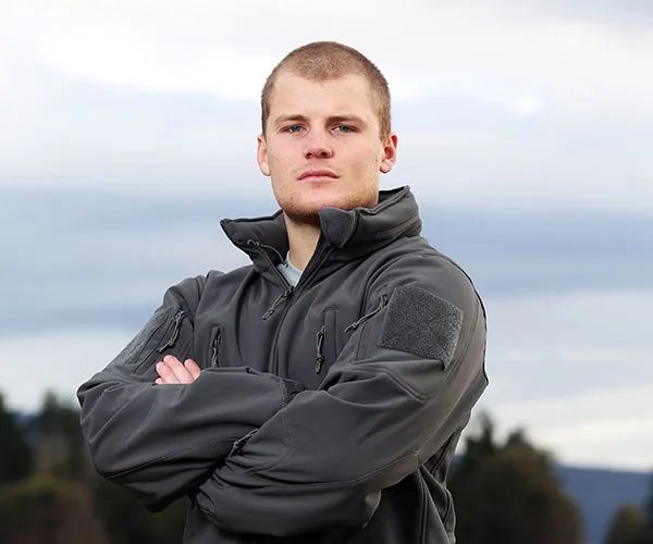 Young man in a tactical jacket with arms crossed, standing outdoors against a cloudy sky.