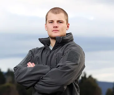 Young man in a tactical jacket with arms crossed, standing outdoors against a cloudy sky.